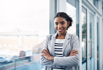 Happy, confident and professional young business woman folding her arms while standing in a modern office. Portrait of a black woman leader smiling, looking proud, powerful, ready and focused