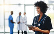© Delmaine Donson/peopleimages.com - Young smiling medical professional doctor, holding digital tablet in modern hospital. Female healthcare specialist standing with test results, with colleagues in background.