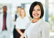 © Daniel Laflor/peopleimages.com - Young, smart and smiling businesswoman with a diverse team at work ready to achieve success. Confident female professional executive standing in a modern office with her colleagues in the background