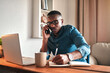 © C Malambo/peopleimages.com - Young serious professional businessman talking on phone call, writing and sitting with digital laptop at home office. African american employee working and consulting in new company startup