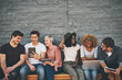 © Camerene Pendl/peopleimages.com - Group of friends on digital devices and social media sitting together while smiling and talking. Technology obsessed online remote workers taking a break. Happy casual creative team outside