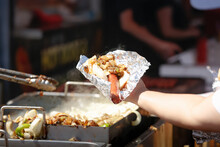 A View Of A Customer Holding Out A Carnival Hot Dog, Waiting For The Vendor To Provide Grilled Toppings.