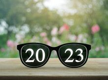 2023 White Text With Black Eye Glasses On Wooden Table Over Blur Pink Flower And Tree In Garden, Business Vision Happy New Year 2023 Cover Concept