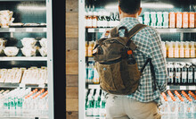 Traveler Backpack Man In Front Of A Drinks And Food Window Store. People And Travel Lifestyle Buying Some Drinks And Sandwiches Before Start The Trip. Concept Of Automatic Machine Shop Station