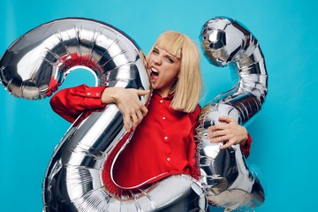 a happy, enthusiastic woman in a red shirt stands on a blue background and holds inflatable balloons in the shape of the number twenty-two in silver color making a funny face expressing her emotions