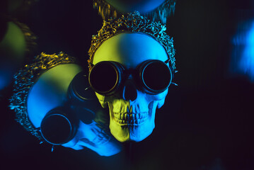 human skull in a crown and steampunk glasses with colored yellow blue neon light on a black background with a mirror reflection. Concept of celebrating a holiday of Dia De Los Muertos