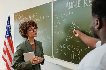 Wall Mural - Young serious female teacher of geometry listening to African American student with piece of chalk pointing at triangle on blackboard