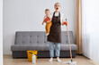 Horizontal shot of little baby girl in young mother hands, desperate woman cleaning at home, tired exhausted mom mopping floor, tidying together in living room.
