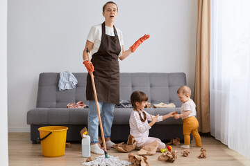 Wall Mural - Portrait of tired mother wearing brown apron cleaning at home, woman with mop cleaning with kids sitting on floor at the sofa, exhausted housewife of tidying up house.