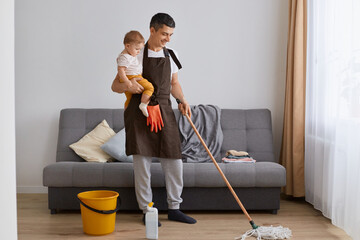 Wall Mural - Portrait of handsome man wearing casual attire and brown apron cleaning house with mop and holding toddler daughter in hands, doing housework, washing floor.