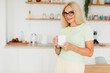 © anatoliycherkas - Modern attractive middle aged woman holding white cup mockup in kitchen at home
