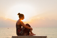 A Young Woman Sits Barefoot On The Oceanfront Bench And Waits For The Sunrise On A Warm Summer Morning. The Orange Light Of The Sunrise Outlines Her Silhouette.