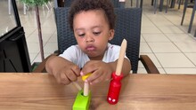 Funny And Happy Two Year Old Afro European Child Playing With Some Wood Toy Tools Seated On A Table.