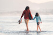 Latino man and his daughter walk barefoot on the beach holding hands, back view