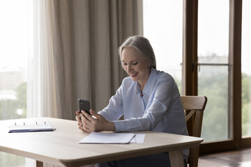 Wall Mural - Happy elderly retired smartphone user talking on video call, enjoying online conversation, holding mobile phone over table with papers at home, smiling, laughing. Retirement, communication concept