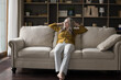 Thoughtful peaceful pretty elderly woman resting on soft couch in living room interior, leaning on back, touching head, relaxing, thinking, dreaming. Home leisure concept. Full length shot