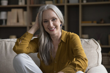 Happy joyful beautiful older 50s woman sitting on couch at home, looking at camera, laughing, smiling, touching long grey hair. Happy retirement, elderly age concept. Head shot portrait,