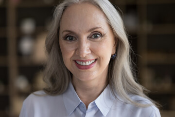 Happy beautiful senior middle aged lady looking at camera with toothy smile, posing indoors. Older business woman, teacher, elderly beauty care model head shot portrait