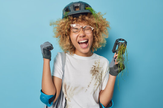 Happy joyful woman clenches fists celebrates success holds bicycle pedal takes break after riding dressed in t shirt and protective gear isolated over blue background. People and active rest concept