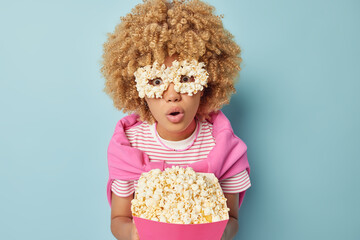 Wall Mural - Indoor shot of shocked amazed curly woman holds popcorn bucket makes eyemask from food watches horror film wears striped t shirt and pink jumper tied over shoulders isolated over blue background