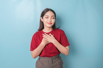 Happy mindful thankful young woman holding hands on chest smiling isolated on blue background feeling no stress, gratitude, mental health balance, peace of mind concept.