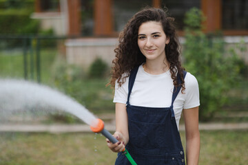 Wall Mural - Young woman with curly hair wearing overall watering plants in country house backyard, garden care, eco-friendly lifestyle, suburban life concept.