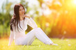 Young woman relax in the early autumn park on green grass. Beauty nature scene with colorful background, trees at fall season. Outdoor lifestyle. Happy smiling woman sitting on green grass