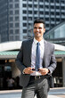© Atstock Productions - Portrait of smiling elegant young Indian businessman wearing gray suit and holding sunglasses while standing against buildings in city