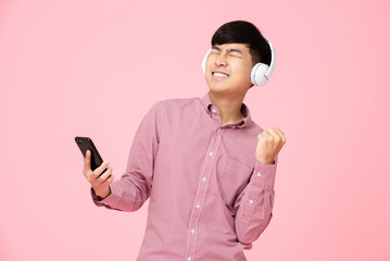 Ecstatic Asian man wearing headphones listening to online streaming music from mobile phone with his fist up in studio pink background