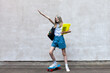 Teen schoolgirl with skateboard,  backpack and document folder raising a hand on a grey wall background. Active blond girl with colored hair strands. Back to school. Copy space. Selective focus.