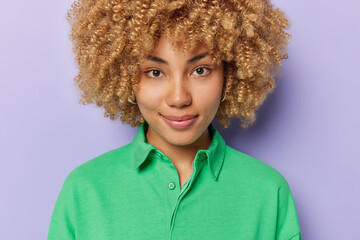 Wall Mural - Portrait of good looking European woman with curly bushy hair dressed in green jumper looks directly at camera has calm expression poses against purple background listens information attentively.
