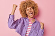 Excited cheerful woman with curly hair clenchs fists celebrates special occasion exclaims from happiness dressed in purple jacket poses against pink background achieves goal or job promotion