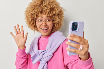 Wall Mural - Positive good looking woman with curly blonde hair waves hello at front camera makes video call records blog talks to best friend abroad says hi smiles friendly wears casual outfit poses indoor.