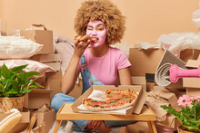 Photo Of Curly Haired European Woman Applies Facial Beauty Mask Eats Appetizing Pizza Enjoys Delicious Snack Moves In New Place For Living Surrounded By Cardboard Boxes Full Of Personal Stuff