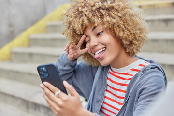 Wall Mural - positive curly haired woman sticks out tongue winks eye makes selfe via smartphone poses outdoors on