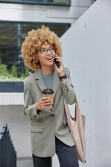 Wall Mural - Outdoor shot of positive curly haired woman entrepreneur has telephone talk drinks takeaway coffee carries bag poses on stairs looks gladfully above enjoys pleasant conversation being on way to work