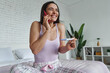 © gstockstudio - Excited woman holding pregnancy test and talking on mobile phone while sitting on bed