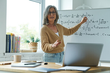 Mature woman teaching mathematics while pointing whiteboard and looking at laptop