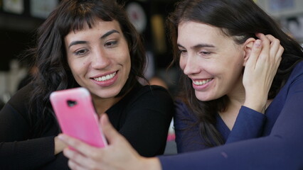 Wall Mural - Two young women looking at smartphone device screen smiling. Female friends sharing information watching content online