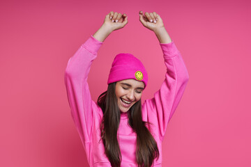 Excited young woman in hooded shirt and funky hat dancing against pink background