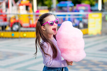 A Child Girl In An Amusement Park In The Summer Eats Cotton Candy Near The Carousels In Sunglasses, The Concept Of Summer Holidays And School Holidays