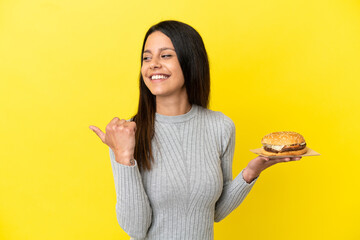 Wall Mural - Young caucasian woman holding a burger isolated on yellow background pointing to the side to present a product