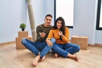 Canvas Print - Young couple sitting on the floor at new home beckoning come here gesture with hand inviting welcoming happy and smiling