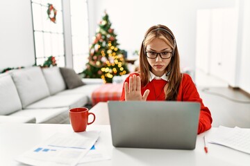 Sticker - Young caucasian girl sitting on the table working using laptop by christmas tree doing stop sing with palm of the hand. warning expression with negative and serious gesture on the face.