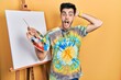 © Krakenimages.com - Young hispanic man standing drawing with palette by painter easel stand crazy and scared with hands on head, afraid and surprised of shock with open mouth