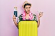© Krakenimages.com - Young hispanic man wearing summer style and hawaiian lei holding passport and plane toy in shock face, looking skeptical and sarcastic, surprised with open mouth