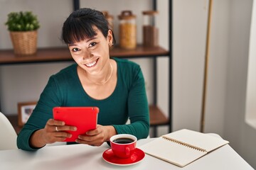 Young beautiful hispanic woman using touchpad drinking coffee sitting on table at home