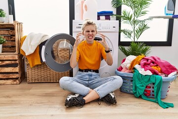 Canvas Print - Young blonde woman doing laundry sitting by washing machine shouting frustrated with rage, hands trying to strangle, yelling mad