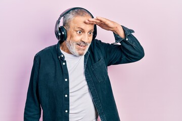 Handsome senior man with beard listening to music using headphones very happy and smiling looking far away with hand over head. searching concept.