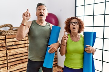 Poster - Middle age couple holding yoga mat amazed and surprised looking up and pointing with fingers and raised arms.